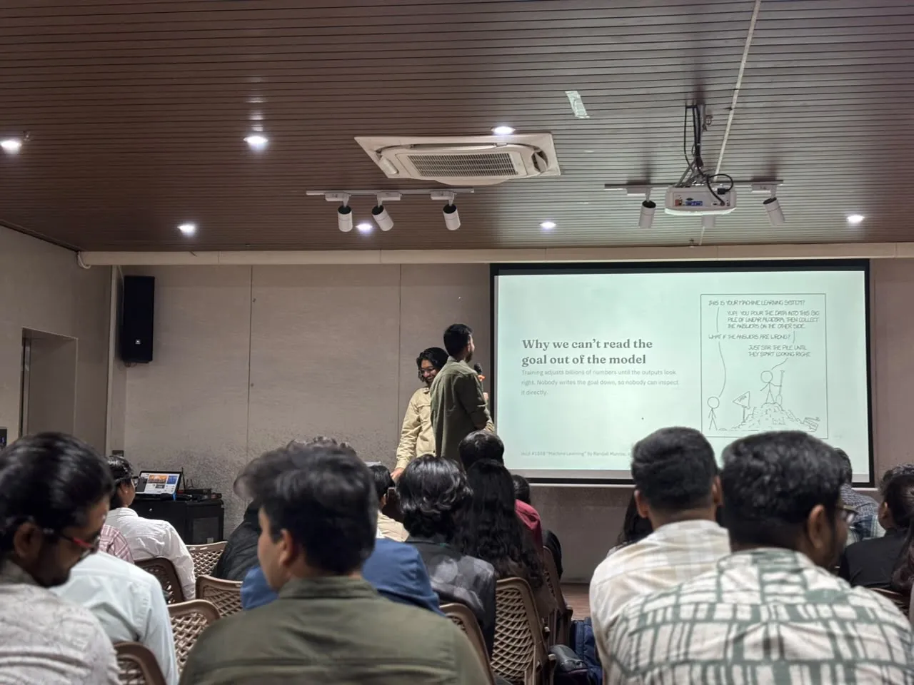 Presenting a slide titled "Why we can't read the goal out of the model", next to an xkcd comic about machine learning